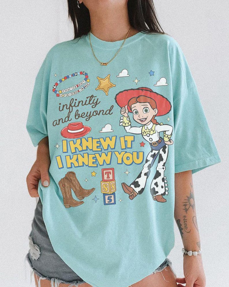May include: A light blue t-shirt featuring a graphic print of the character Jessie from Toy Story, brown cowboy boots, a red hat, a yellow star, and colorful blocks. The text reads infinity and beyond, I KNEW IT, I KNEW YOU, and YOU'VE GOT A FRIEND IN ME.