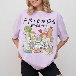 K&ouml;nnte beinhalten: Helllila T-Shirt mit einer Grafik von Toy Story-Charakteren, darunter Woody und Buzz Lightyear. Das Wort "FRIENDS" ist &uuml;ber den Charakteren aufgedruckt, darunter "SINCE 1995". Das Design ist eine Anspielung auf die Fernsehserie Friends.