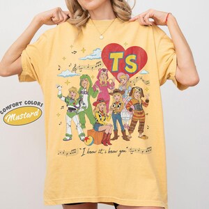 Puede incluir: Una camiseta de manga corta color amarillo mostaza con un estampado gr&aacute;fico de personajes disfrazados, un coraz&oacute;n rojo con las letras TS, notas musicales, nubes y el texto "I knew it, i knew you" impreso en la parte inferior.