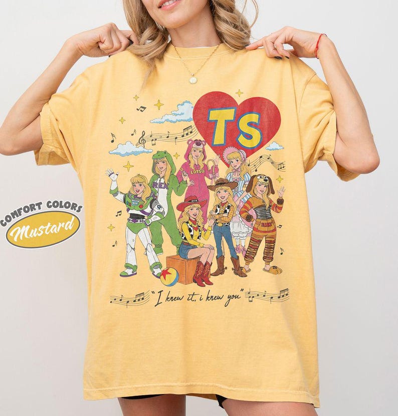 Puede incluir: Una camiseta de manga corta color amarillo mostaza con un estampado gr&aacute;fico de personajes disfrazados, un coraz&oacute;n rojo con las letras TS, notas musicales, nubes y el texto "I knew it, i knew you" impreso en la parte inferior.