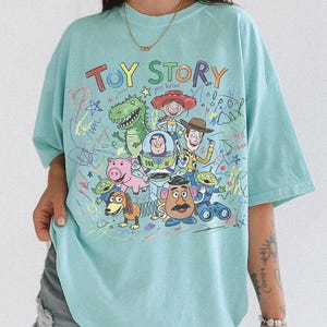 K&ouml;nnte beinhalten: Hellblaues T-Shirt mit dem Schriftzug "TOY STORY" in bunten Buchstaben &uuml;ber einer Grafik von Figuren aus dem Zeichentrickfilm. Das Design umfasst Woody, Buzz Lightyear und andere Charaktere mit bunten Kritzeleien.