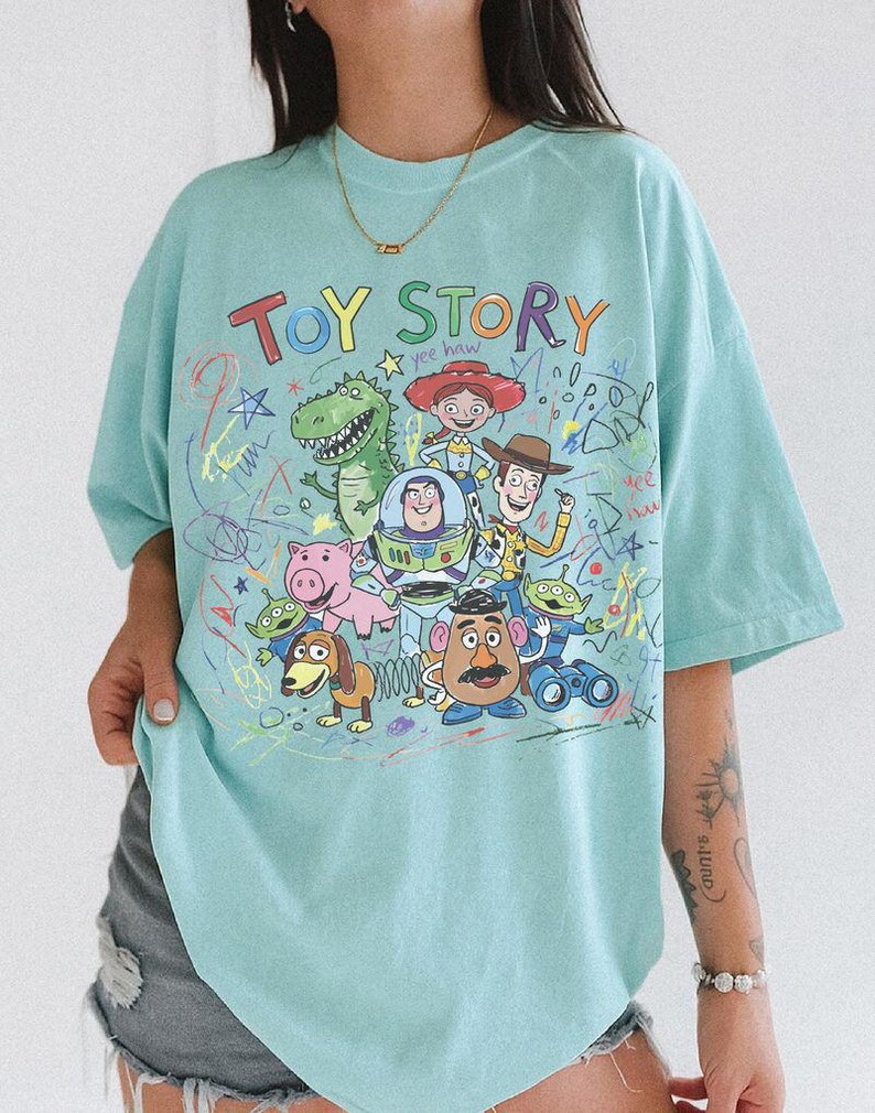 K&ouml;nnte beinhalten: Hellblaues T-Shirt mit dem Schriftzug "TOY STORY" in bunten Buchstaben &uuml;ber einer Grafik von Figuren aus dem Zeichentrickfilm. Das Design umfasst Woody, Buzz Lightyear und andere Charaktere mit bunten Kritzeleien.