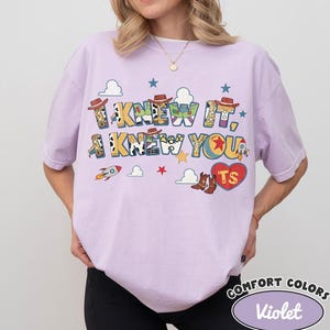 May include: A light purple short-sleeve t-shirt featuring a graphic print with the text I KNEW IT, I KNEW YOU and TS inside a red heart. The design includes cowboy hats, boots, rockets, stars, and clouds in various colors and patterns.