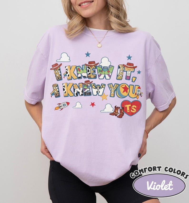 May include: A light purple short-sleeve t-shirt featuring a graphic print with the text I KNEW IT, I KNEW YOU and TS inside a red heart. The design includes cowboy hats, boots, rockets, stars, and clouds in various colors and patterns.