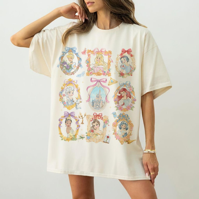 May include: Cream-colored t-shirt featuring nine framed portraits of animated princesses. Each portrait is surrounded by a decorative gold frame with floral and bow accents. The portraits include various princesses in their signature attire.