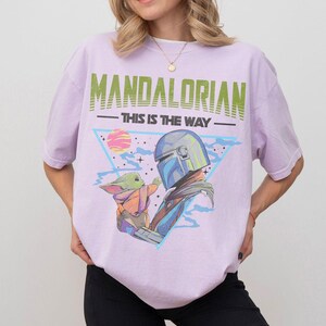 Może przedstawiać: Jasnofioletowy t-shirt z nadrukiem graficznym przedstawiającym Mandalorianina i Grogu. Tekst głosi MANDALORIAN oraz THIS IS THE WAY. Wz&oacute;r zawiera niebieski tr&oacute;jkąt, r&oacute;żową planetę i gwiazdy na jasnofioletowym materiale.