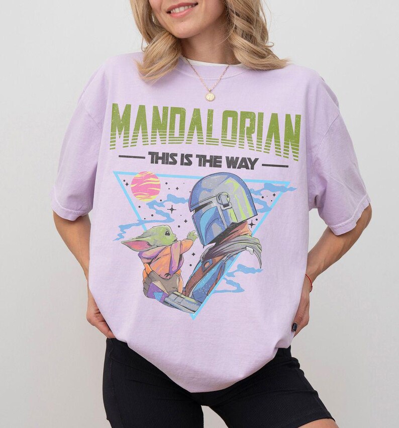 Może przedstawiać: Jasnofioletowy t-shirt z nadrukiem graficznym przedstawiającym Mandalorianina i Grogu. Tekst głosi MANDALORIAN oraz THIS IS THE WAY. Wz&oacute;r zawiera niebieski tr&oacute;jkąt, r&oacute;żową planetę i gwiazdy na jasnofioletowym materiale.