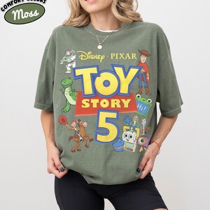 Puede incluir: Una camiseta de manga corta color verde musgo con un estampado gr&aacute;fico que incluye el texto Disney Pixar Toy Story 5. El dise&ntilde;o presenta ilustraciones de Buzz Lightyear, Woody, Rex, Jessie sobre Perdig&oacute;n, Forky y otros personajes en amarillo, azul y rojo.
