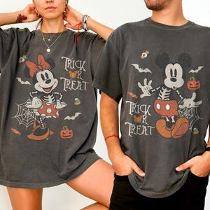 May include: Two charcoal gray t-shirts featuring vintage-style Halloween graphics. One shirt displays Minnie Mouse in a skeleton costume with the text Trick or Treat, while the other shows Mickey Mouse in a skeleton costume with the same text.