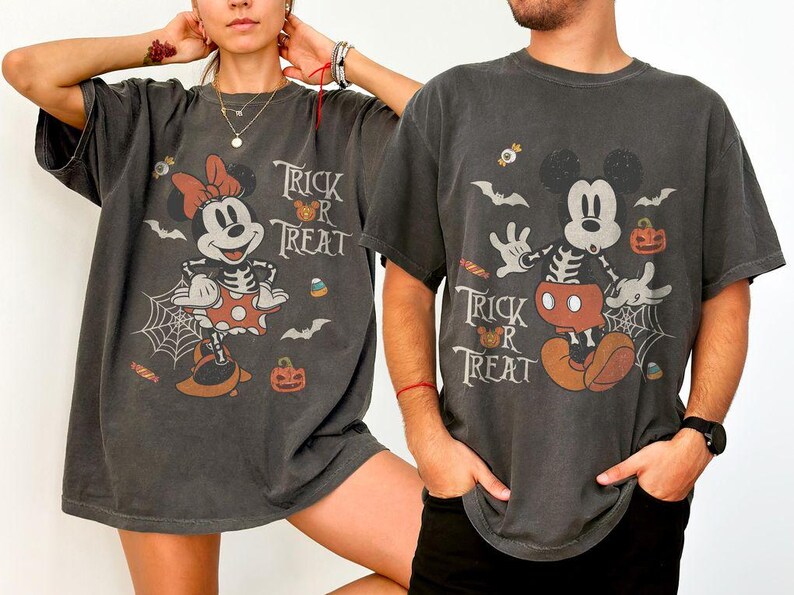May include: Two charcoal gray t-shirts featuring vintage-style Halloween graphics. One shirt displays Minnie Mouse in a skeleton costume with the text Trick or Treat, while the other shows Mickey Mouse in a skeleton costume with the same text.