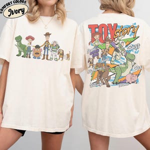 Może przedstawiać: Kremowa koszulka z kolorową grafiką postaci z Toy Story, w tym Chudego i Buzz'a Astrala. Prz&oacute;d koszulki przedstawia postacie, a tył logo Toy Story i więcej postaci. Koszulka wykonana jest z miękkiego, wygodnego materiału.
