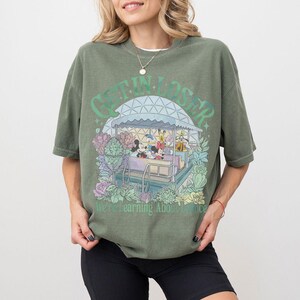Pode incluir: Camiseta verde-oliva com um gr&aacute;fico de personagens da Disney em um passeio. O texto "GET IN LOSER" est&aacute; acima do passeio, com "We're Learning About Balance" abaixo. O design &eacute; cercado por flores e folhagens em tons pastel.