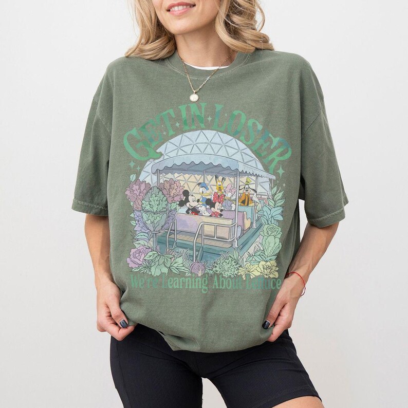 Pode incluir: Camiseta verde-oliva com um gr&aacute;fico de personagens da Disney em um passeio. O texto "GET IN LOSER" est&aacute; acima do passeio, com "We're Learning About Balance" abaixo. O design &eacute; cercado por flores e folhagens em tons pastel.