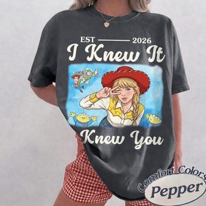 May include: A charcoal gray t-shirt featuring a graphic print of a blonde character wearing a red cowboy hat, a yellow shirt, and making a peace sign. The design includes the text EST 2026, I Knew It, and Knew You, along with small cartoon figures of a cowboy, a space ranger, and green aliens.