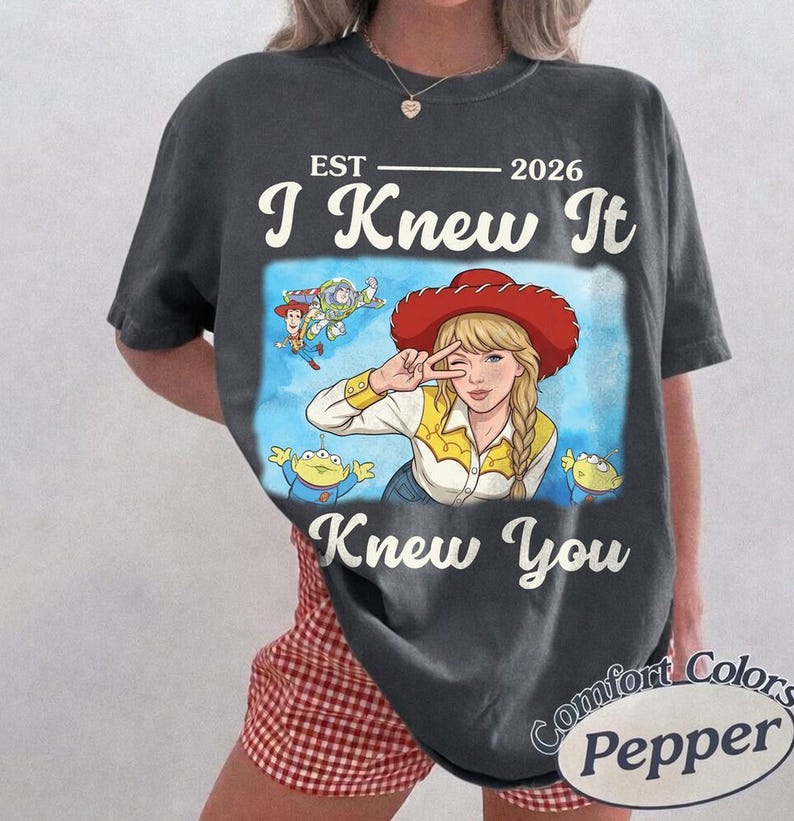 May include: A charcoal gray t-shirt featuring a graphic print of a blonde character wearing a red cowboy hat, a yellow shirt, and making a peace sign. The design includes the text EST 2026, I Knew It, and Knew You, along with small cartoon figures of a cowboy, a space ranger, and green aliens.