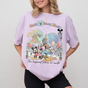 May include: A light purple t-shirt featuring a colorful graphic of Disney characters and landmarks. The design includes Mickey and Minnie Mouse, Goofy, Donald and Daisy Duck, and the text "Walt Disney World" and "The happiest place on earth."