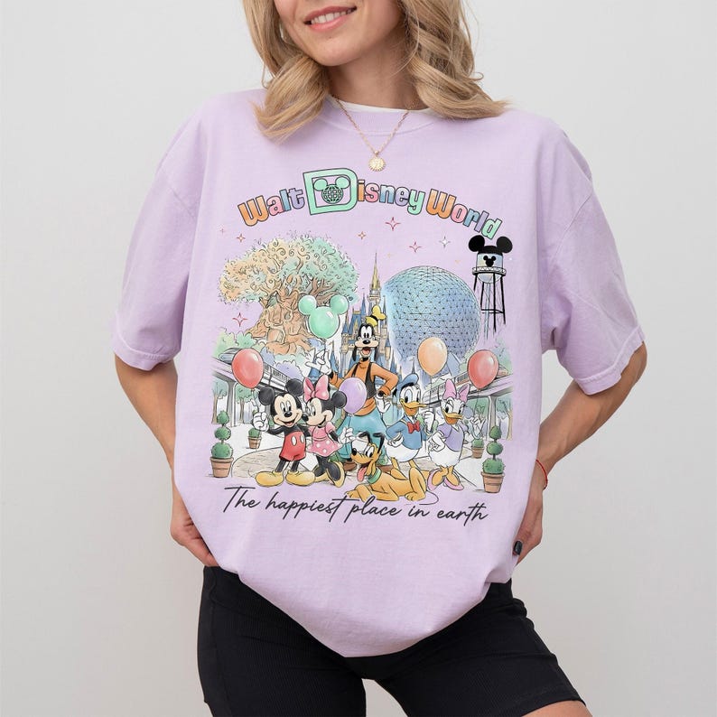 May include: A light purple t-shirt featuring a colorful graphic of Disney characters and landmarks. The design includes Mickey and Minnie Mouse, Goofy, Donald and Daisy Duck, and the text "Walt Disney World" and "The happiest place on earth."
