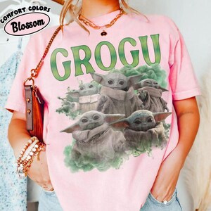 May include: A light pink short-sleeve t-shirt featuring a graphic print of four Grogu characters surrounded by green smoke. The text GROGU is printed in large green letters above the characters. A small logo in the corner reads COMFORT COLORS Blossom.