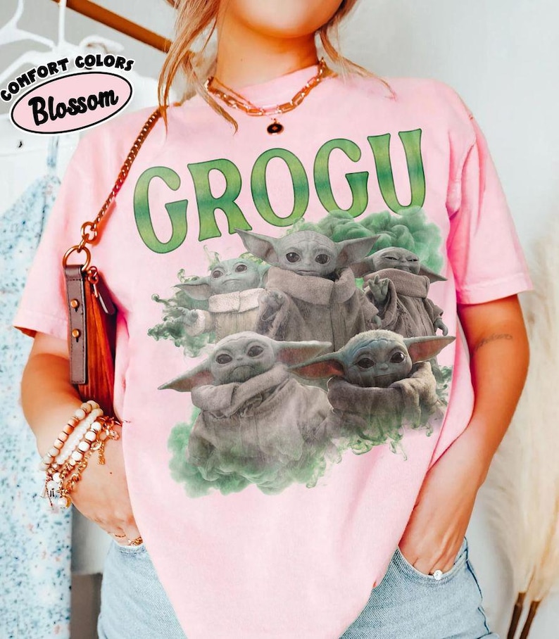 May include: A light pink short-sleeve t-shirt featuring a graphic print of four Grogu characters surrounded by green smoke. The text GROGU is printed in large green letters above the characters. A small logo in the corner reads COMFORT COLORS Blossom.