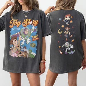 May include: Dark gray t-shirt featuring a colorful Toy Story graphic. The front of the shirt displays Buzz Lightyear, Woody, and other characters with the text "Toy Story Est. 1995." The back of the shirt has additional character graphics.