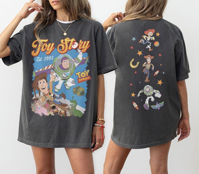May include: Dark gray t-shirt featuring a colorful Toy Story graphic. The front of the shirt displays Buzz Lightyear, Woody, and other characters with the text "Toy Story Est. 1995." The back of the shirt has additional character graphics.