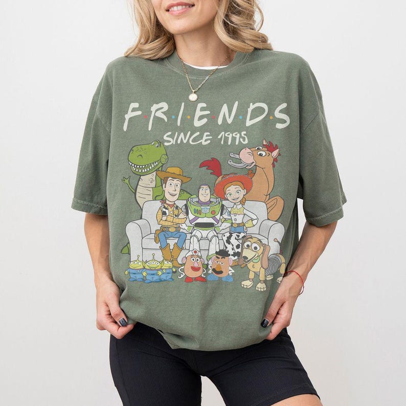 K&ouml;nnte beinhalten: Olivgr&uuml;nes T-Shirt mit dem Aufdruck "FRIENDS" und dem Text "SINCE 1995". Das T-Shirt zeigt eine Grafik von Toy Story-Charakteren. Das T-Shirt hat einen Rundhalsausschnitt und kurze &Auml;rmel.