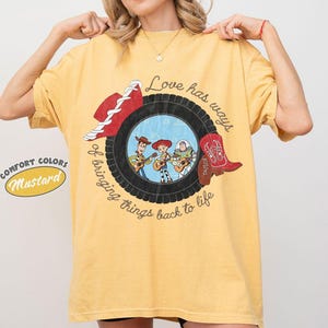 May include: A mustard yellow t-shirt featuring a graphic of a black tire surrounding a scene with three cartoon characters playing guitars. The text reads, Love has ways of bringing things back to life. A red cowboy hat and a red cowboy boot are included.