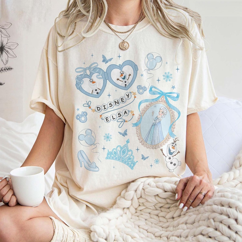 May include: Cream-colored t-shirt featuring a Disney Elsa design. The design includes images of Elsa, Olaf, and other Frozen-themed elements in shades of blue. The text "Disney Elsa" is also present.