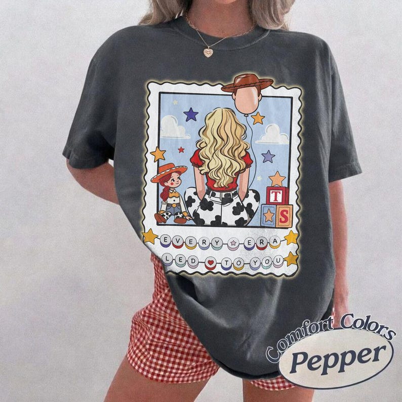 May include: A charcoal gray t-shirt featuring a graphic print of a blonde person seen from behind, a small cowgirl doll, a cowboy hat balloon, and alphabet blocks. The text reads EVERY ERA LED TO YOU, surrounded by stars on a light blue background.