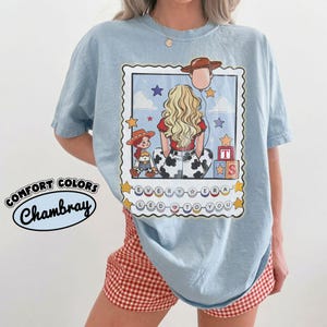May include: A light blue t-shirt featuring a graphic print of a blonde person seen from behind, wearing a red top and cow-print pants. The design includes a cowboy hat balloon, a small doll, alphabet blocks, and the text "EVERY ERA LED TO YOU".