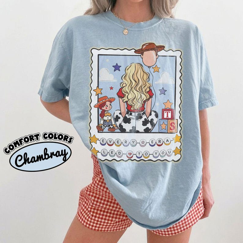 May include: A light blue t-shirt featuring a graphic print of a blonde person seen from behind, wearing a red top and cow-print pants. The design includes a cowboy hat balloon, a small doll, alphabet blocks, and the text "EVERY ERA LED TO YOU".