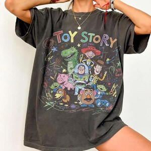 K&ouml;nnte beinhalten: Dunkelgraues T-Shirt mit einer farbenfrohen Grafik von Charakteren aus dem Animationsfilm "Toy Story". Das Design enth&auml;lt die Worte "Toy Story" in einer Regenbogen-Schriftart sowie Bilder von Woody, Buzz Lightyear und anderen Charakteren.