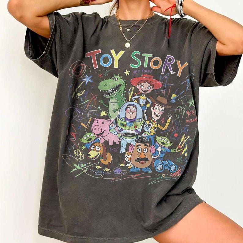 K&ouml;nnte beinhalten: Dunkelgraues T-Shirt mit einer farbenfrohen Grafik von Charakteren aus dem Animationsfilm "Toy Story". Das Design enth&auml;lt die Worte "Toy Story" in einer Regenbogen-Schriftart sowie Bilder von Woody, Buzz Lightyear und anderen Charakteren.