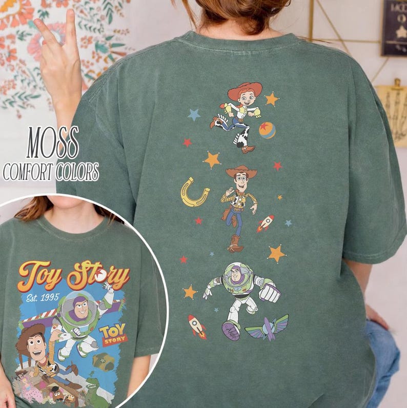 May include: Moss green t-shirt featuring characters from the animated film "Toy Story." The front of the shirt displays Buzz Lightyear, Woody, and other characters. The back of the shirt has Jessie, Woody, and Buzz Lightyear graphics. The text "Toy Story Est. 1995" is also visible.