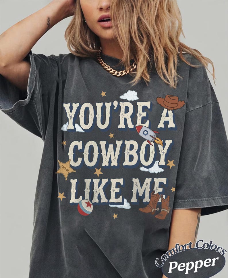 May include: A charcoal gray t-shirt featuring the text "YOU'RE A COWBOY LIKE ME" in a distressed font. The graphic includes a brown cowboy hat, a rocket ship, a sheriff star badge labeled "TAYLOR", a red and white ball, and brown cowboy boots.