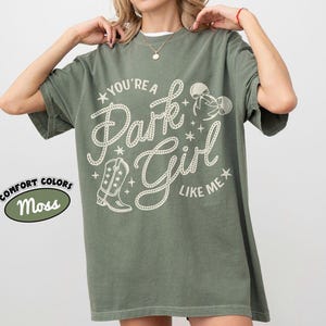 Op de afbeelding: Een mosgroen T-shirt met korte mouwen en een cr&egrave;mekleurig grafisch ontwerp met de tekst "YOU'RE A Park Girl LIKE ME". Het ontwerp bevat een cowboylaars, een strik en sterren. Een klein ovaal label geeft de kleur Moss aan.
