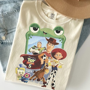 May include: A cream-colored t-shirt featuring a graphic of Toy Story characters including Woody, Jessie, Bullseye, and Buzz Lightyear framed by a green frog outline. The text reads Disney Pixar Toy Story 5 In Theaters June 19.