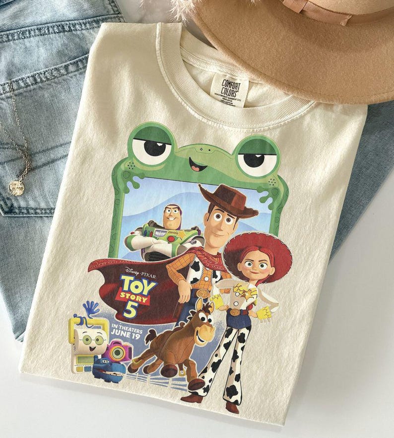 May include: A cream-colored t-shirt featuring a graphic of Toy Story characters including Woody, Jessie, Bullseye, and Buzz Lightyear framed by a green frog outline. The text reads Disney Pixar Toy Story 5 In Theaters June 19.
