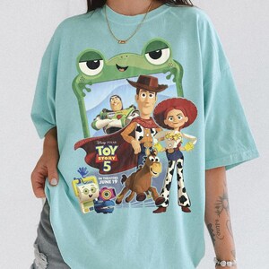 May include: A light blue t-shirt featuring a graphic print of Toy Story 5 characters including Woody, Jessie, Buzz Lightyear, and Bullseye. The design is framed by a green frog-shaped border and includes the text Toy Story 5 In Theaters June 19.