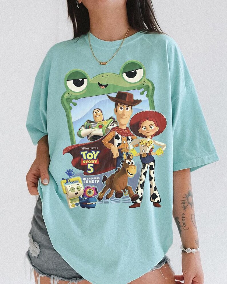 May include: A light blue t-shirt featuring a graphic print of Toy Story 5 characters including Woody, Jessie, Buzz Lightyear, and Bullseye. The design is framed by a green frog-shaped border and includes the text Toy Story 5 In Theaters June 19.
