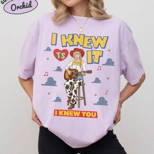 May include: A light purple short-sleeve t-shirt featuring a graphic of a person in a cowboy hat and cow-print pants playing an acoustic guitar on a stool. The text reads I KNEW TS IT and I KNEW YOU, with clouds, stars, and musical notes.