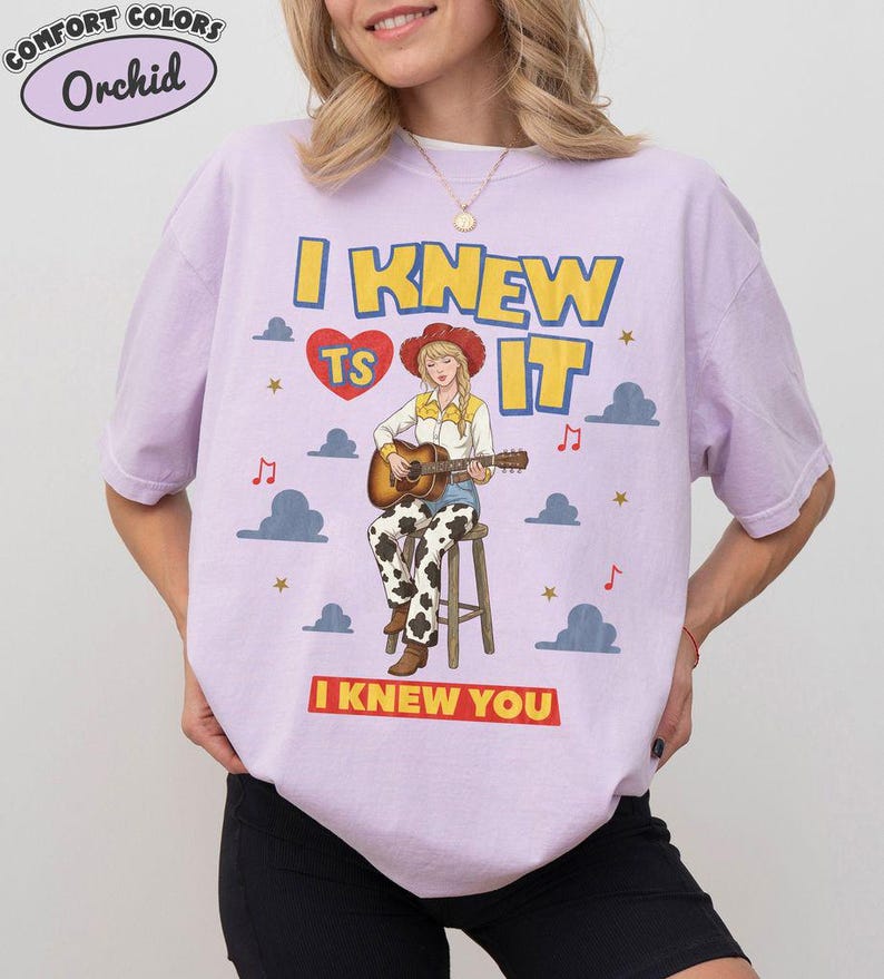 May include: A light purple short-sleeve t-shirt featuring a graphic of a person in a cowboy hat and cow-print pants playing an acoustic guitar on a stool. The text reads I KNEW TS IT and I KNEW YOU, with clouds, stars, and musical notes.