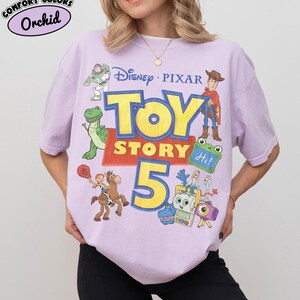 Puede incluir: Una camiseta de color morado claro con un estampado gr&aacute;fico que incluye el texto Disney Pixar Toy Story 5. El dise&ntilde;o muestra ilustraciones de Buzz Lightyear, Woody, Rex, Jessie sobre Perdig&oacute;n y otros personajes. Una etiqueta indica Comfort Colors Orchid.