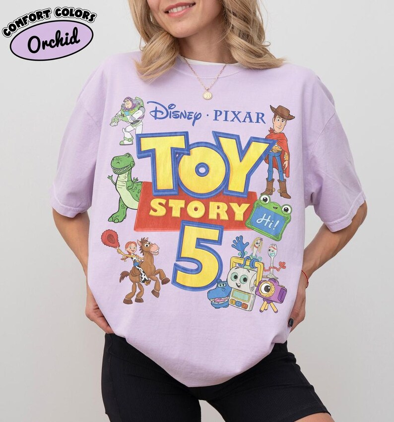 Puede incluir: Una camiseta de color morado claro con un estampado gr&aacute;fico que incluye el texto Disney Pixar Toy Story 5. El dise&ntilde;o muestra ilustraciones de Buzz Lightyear, Woody, Rex, Jessie sobre Perdig&oacute;n y otros personajes. Una etiqueta indica Comfort Colors Orchid.