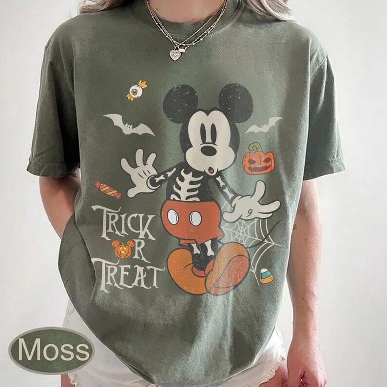 May include: A moss green short-sleeve t-shirt featuring a graphic of Mickey Mouse with a skeleton ribcage design. The print includes bats, a pumpkin, candy, a spiderweb, and the text "TRICK OR TREAT" with a small pumpkin icon.