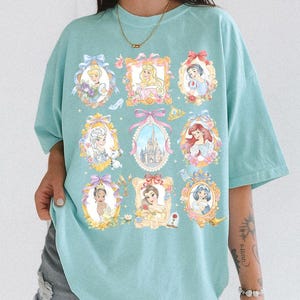 May include: A light blue t-shirt featuring framed portraits of various princesses. Each portrait is surrounded by floral and bow accents. The shirt has a relaxed fit and short sleeves, perfect for casual wear.