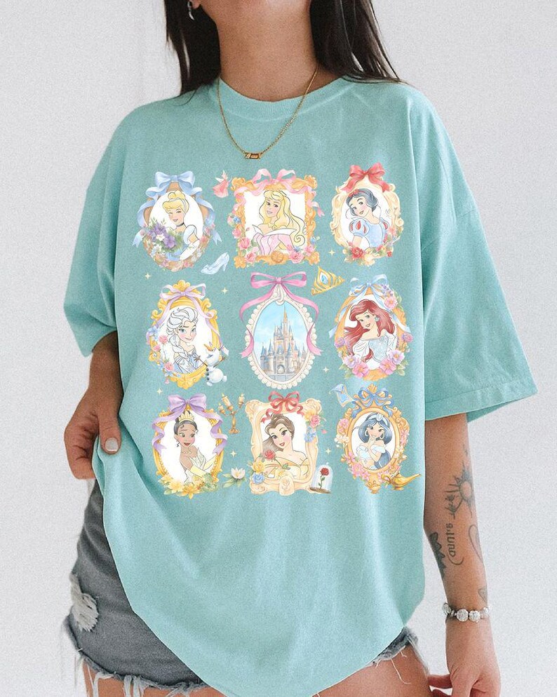 May include: A light blue t-shirt featuring framed portraits of various princesses. Each portrait is surrounded by floral and bow accents. The shirt has a relaxed fit and short sleeves, perfect for casual wear.