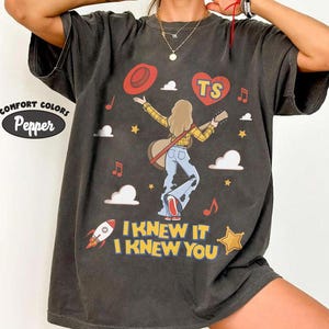 Puede incluir: Una camiseta gris carb&oacute;n con un gr&aacute;fico de una persona de pelo largo tocando la guitarra, vistiendo una camisa de cuadros amarilla y vaqueros azules. El dise&ntilde;o incluye un sombrero rojo, un coraz&oacute;n rojo con el texto TS, nubes, estrellas, un cohete y el texto I KNEW IT I KNEW YOU.