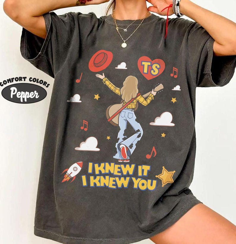 Puede incluir: Una camiseta gris carb&oacute;n con un gr&aacute;fico de una persona de pelo largo tocando la guitarra, vistiendo una camisa de cuadros amarilla y vaqueros azules. El dise&ntilde;o incluye un sombrero rojo, un coraz&oacute;n rojo con el texto TS, nubes, estrellas, un cohete y el texto I KNEW IT I KNEW YOU.