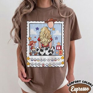 May include: A brown t-shirt featuring a graphic print of a person with long blonde hair, a cowgirl doll, and a cowboy hat balloon. The design includes alphabet blocks, stars, clouds, and the text "EVERY ERA LED TO YOU" on circular beads.